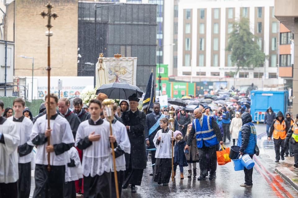 Rosary Crusade procession in the rain, led by clergy and altar servers carrying a statue of Our Lady of Fatima